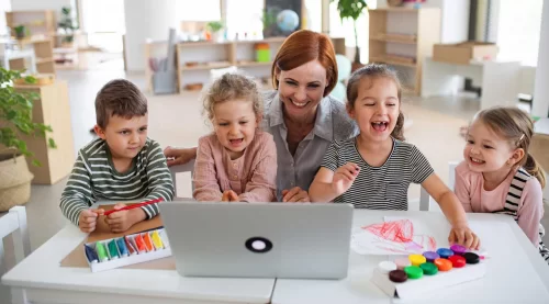 Teacher laughing with five preschoolers around a laptop, paints and modeling clay