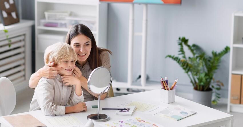 Smiling caregiver hugging a young boy at a table with a mirror, workbooks and colored pencils