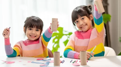 Two smiling girls in striped sweaters holding up cardboard tubes at a craft table