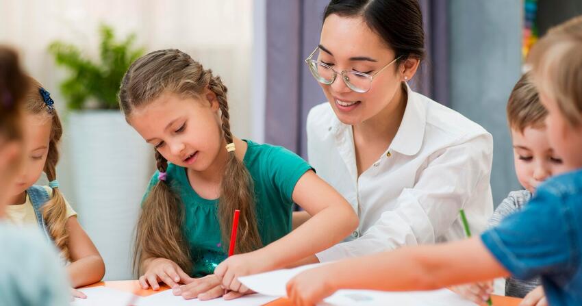 Teacher in glasses helping a girl in braids draw at a table with other young children
